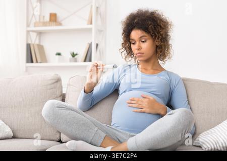 Sad mixed race pregnant woman checking body temperature sitting on the sofa at home Stock Photo