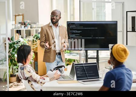 Black male programmer wearing eyeglasses discussing codes while standing in front of colleagues in office Stock Photo