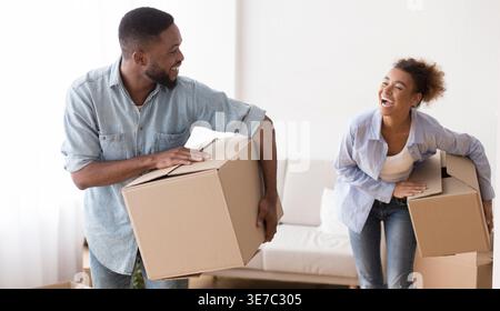 Cheerful Black couple looking at each other laughing carrying moving boxes walking in new home Stock Photo