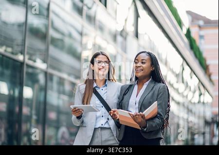 Smiling Black businesswoman with braids holding book talking to caucasian female colleague with tablet walking outside office building during daytime Stock Photo