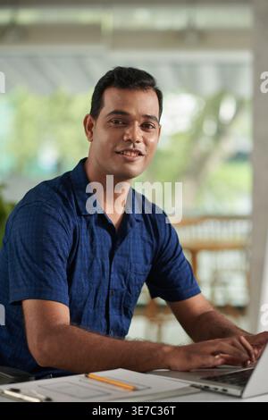 Smiling Indian male designer using laptop sitting at table with clipboard in office Stock Photo