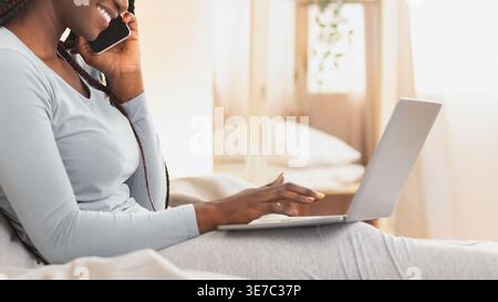 Smiling Black woman with braids wearing pajamas using laptop while having phone conversation sitting on bed in bedroom at home Stock Photo