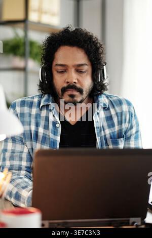 Indian male offie worker with curly hair wearing headphones sitting at desk and working on laptop in office Stock Photo