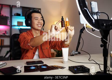 Asian man wearing headphones holding face serum bottles making content for blog while sitting at table facing ring light at home Stock Photo
