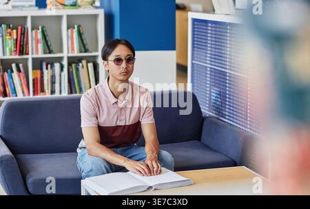 Blind Asian male student wearing sunglasses reading book in tactile braille sitting on couch in college library Stock Photo