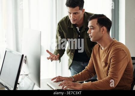 Indian male programmers leaning forward beside Indian male colleague typing codes on computer in office Stock Photo