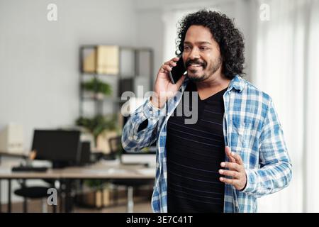 Indian male web developer with curly hair wearing plaid shirt having phone call looking away while standing in office Stock Photo