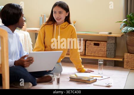 Asian female and Black female students talking and sitting on the floor while doing homework using laptop inside their dorm room Stock Photo