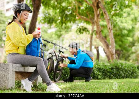 Smiling Asian woman in helmet sitting on bench getting water in backpack near Asian man fixing bike in park during daytime Stock Photo