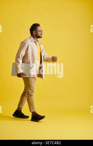 Smiling Asian man with eyeglasses carrying laptop walking against yellow background in studio Stock Photo