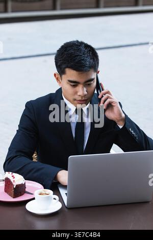 Serious Asian businessman having phone call and working on laptop sitting at table with coffee and slice of cake on plate in outdoor cafe Stock Photo