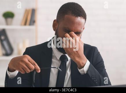 Stressed Black businessman holding eyeglasses touching his nose bridge sitting with closed eyes in office Stock Photo