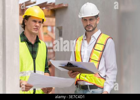 Hispanic male engineer with hardhat and safety vest discussing project with caucasian male colleague in construction site Stock Photo