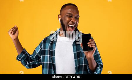 Black man with closed eyes wearing earphones singing using smartphone while standing against yellow background in studio Stock Photo