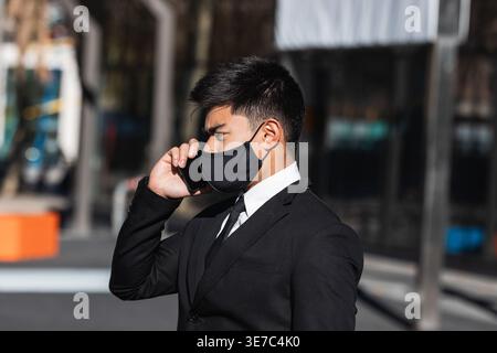 Serious Asian businessman wearing face mask having phone call looking away standing on city street on sunny day Stock Photo