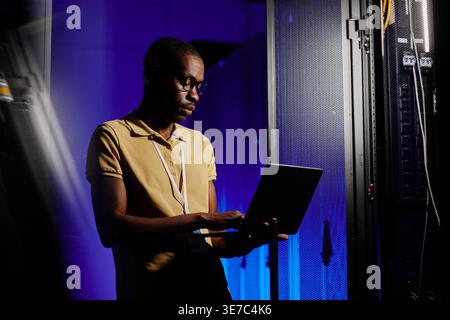Black male IT engineer with eyeglasses using laptop standing in server room with blue neon lights in data center Stock Photo
