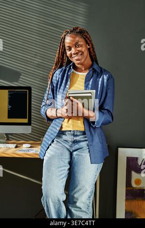 Black woman with braids holding notebooks standing and leaning against table with computer monitor in office Stock Photo