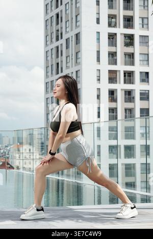 Asian woman in fitness wear doing legs stretching exercise by the swimming pool on rooftop Stock Photo