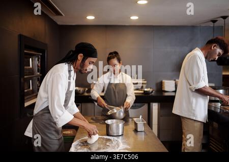 Black male baker making dough standing at table near colleagues in restaurant kitchen Stock Photo