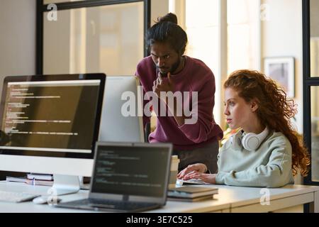 Indian male programmer with hand on chin standing near caucasian female colleague working on computer sitting at table in office Stock Photo