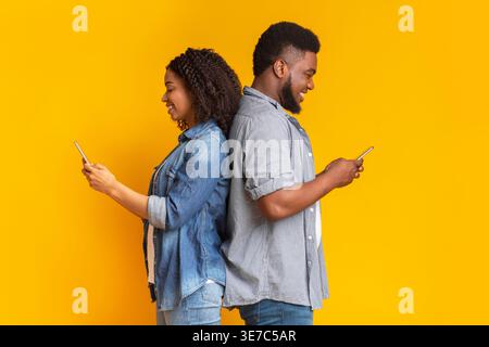Black couple standing back to back while using their smartphone against yellow background Stock Photo