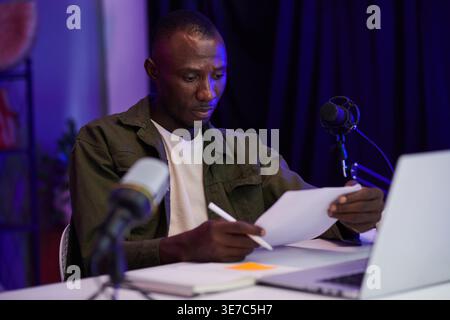 Black male podcaster holding pen and reading his script while sitting at table with laptop and headphones in his home studio Stock Photo