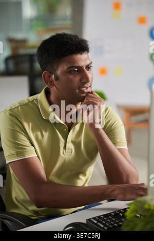 Serious Indian male designer working on computer sitting with hand on chin at table in office Stock Photo