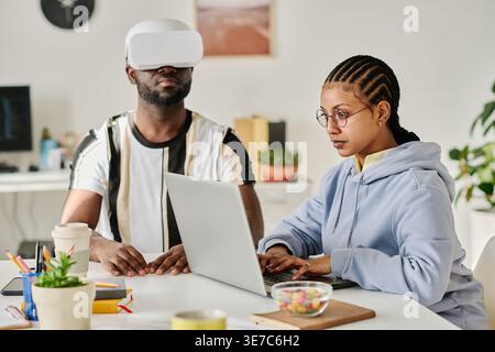 Mixed race woman with braids working on laptop sitting at desk with Black man wearing VR in office Stock Photo