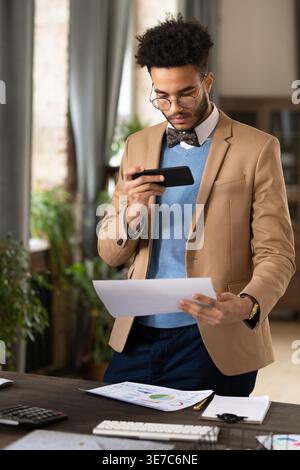 Mixed-race businessman with short afro wearing eyeglasses taking picture of document using smartphone while standing at table with computer and cal... Stock Photo