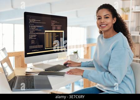 Mixed-race female programmer sitting at table with laptop and computer in office Stock Photo