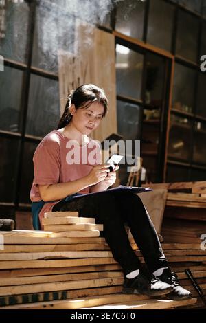 Asian female carpenter using smartphone sitting with clipboard on lap sitting on pile of wooden planks in workshop during daytime Stock Photo