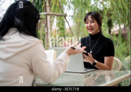 Asian woman working remotely with her colleague while sitting at table in garden during daytime Stock Photo