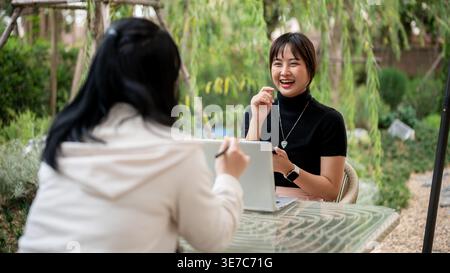 Asian woman working remotely with her colleague while sitting at table in garden during daytime Stock Photo