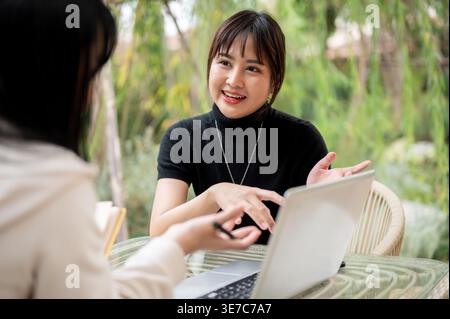 Asian woman working remotely with her colleague while sitting at table in garden during daytime Stock Photo