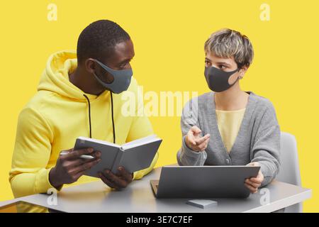 Diverse group of people with laptop and  notebook wearing face masks having conversation while sitting together at table in front of yellow backgro... Stock Photo