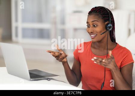 Teleconference Concept. Young Black Woman In Headphones Making Video Call On Laptop While Working At Home Office, Smiling African American Lady Talkin Stock Photo