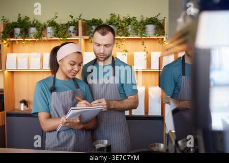 Mixed-race female and caucasian male employees wearing aprons doing inventory while standing at the counter in cafe Stock Photo