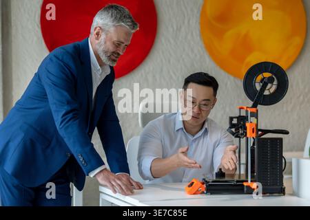 Asian male industrial engineer wearing eyeglasses sitting at table and presenting 3D printer to caucasian male colleague standing beside him in office Stock Photo