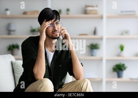 Upset Middle Eastern man wearing eyeglasses having phone call looking away touching head while sitting on sofa in living room at home Stock Photo