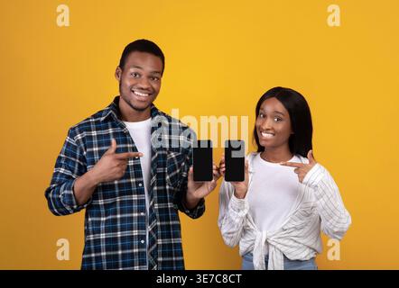 Black couple pointing fingers and showing smartphones while standing against yellow background in studio Stock Photo