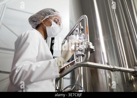 Mixed-race woman worker wearing protective mask and lab coat operating equipment in chemical factory Stock Photo