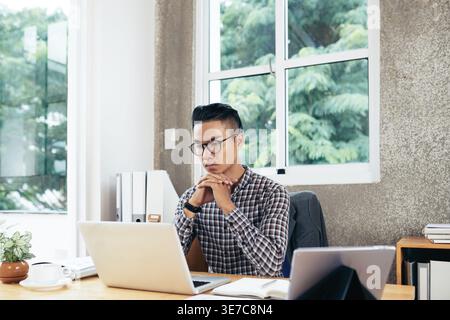 Mixed race man looking at laptop while sitting with clasped hands at table in office Stock Photo