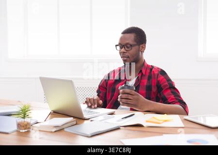 Black businessman wearing eyeglasses holding coffee cup while working on laptop sitting at table in office Stock Photo
