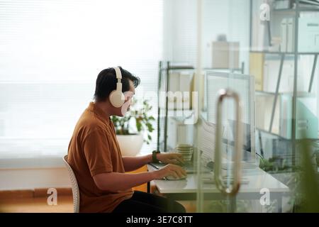 Asian male software developer wearing headphones testing new software on laptop sitting at desk in office Stock Photo