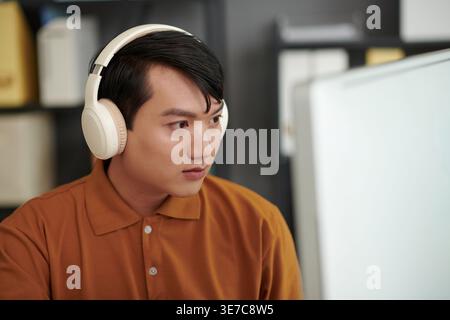 Asian male software developer wearing headphones testing new software on laptop sitting at desk in office Stock Photo
