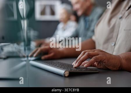 Hands of Black middle-aged woman typing on keyboard sitting beside senior classmates in office Stock Photo