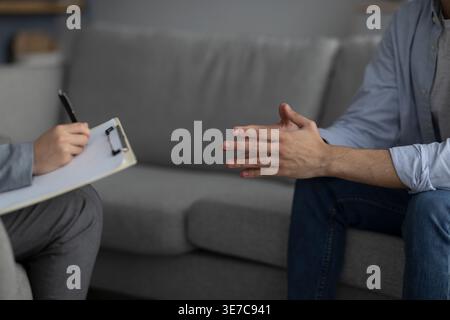 Cropped image of Middle Eastern man with hands together in front of female psychologist taking notes while sitting on couch in clinic Stock Photo