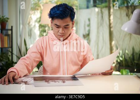 Asian male with blue hair looking and choosing photos while sitting at table at home during daytime Stock Photo