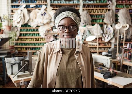 Black female tailor wearing eyeglasses standing against table with sewing machine in workshop Stock Photo
