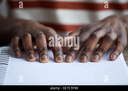 Close-up of hands of blind Black man reading book in braille over the wooden table indoors Stock Photo